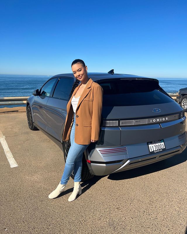 Maurissa Gunn in a brown coat, blue pants, and white boots in front of her Ford Mustang Mach-E car.